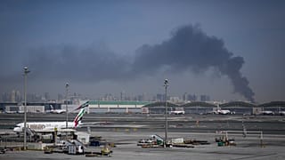 A plume of smoke caused by an Iranian strike is seen in the background as Emirates planes are parked at Dubai International Airport after its closure