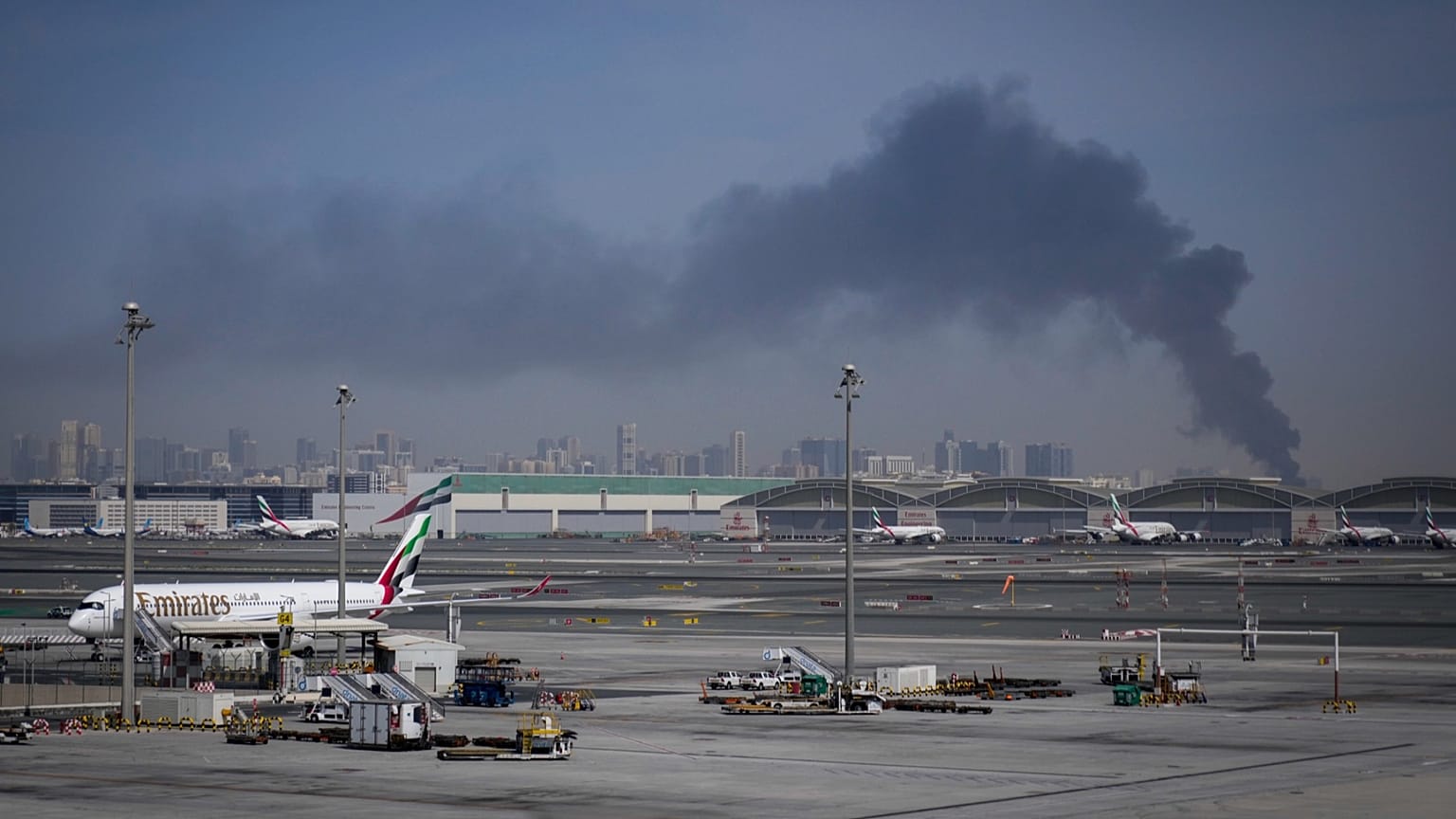 A plume of smoke caused by an Iranian strike is seen in the background as Emirates planes are parked at Dubai International Airport