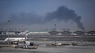A plume of smoke caused by an Iranian strike is seen in the background as Emirates planes are parked at Dubai International Airport