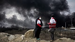 Two women from the Iranian Red Crescent Society stand as a thick plume of smoke from a US-Israeli strike on an oil storage facility in Tehran, Iran, Sunday, March 8, 2026