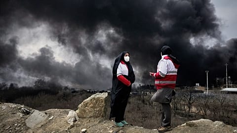 Two women from the Iranian Red Crescent Society stand as a thick plume of smoke from a US-Israeli strike on an oil storage facility in Tehran, Iran, Sunday, March 8, 2026