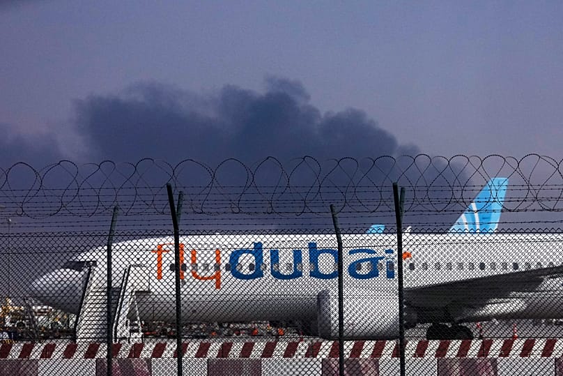 Um avião da FlyDubai está estacionado no Aeroporto Internacional do Dubai enquanto fumo se ergue ao fundo, depois de um drone ter atingido um depósito de combustível na madrugada de 16 de março de 2026.