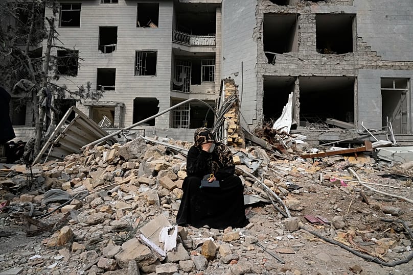 A woman sits on rubble across from a residential building damaged in air strikes on Tehran, 12 March, 2026