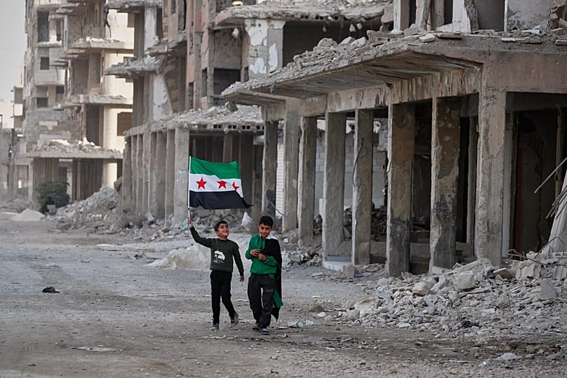 Two boys walk down a street carrying Syrian flags in the war-damaged Damascus suburb of Daraya, 5 December, 2025