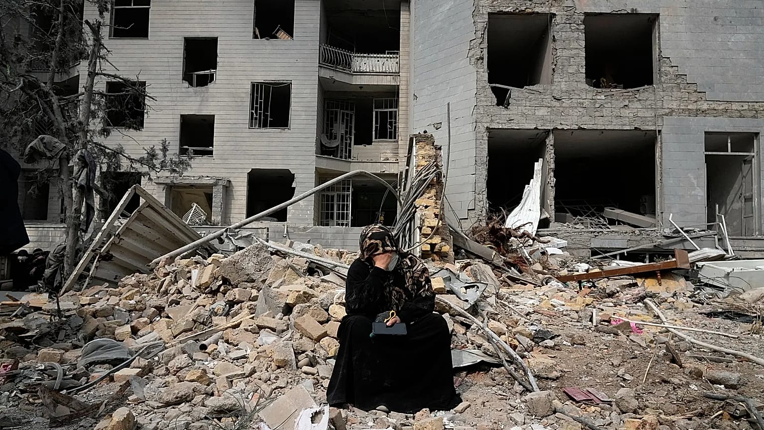 A woman sits on rubble across from a residential building damaged in air strikes in Tehran, 12 March, 2026