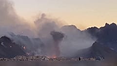 In der Ferne sind Rauch und Explosionen zu sehen, im Vordergrund stehen Häuser.