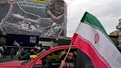 رجل يلوّح بعلامة النصر وهو يحمل العلم الإيراني أمام لوحة إعلانية مناهضة للولايات المتحدة، ميدان الثورة الإسلامية بوسط طهران، إيران، يوم الاثنين 13 أبريل/نيسان 2026. 