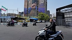 تمر دراجات نارية أمام لوحة إعلانية تظهر رسمًا بيانيًا يصور يد عسكري ممسكًا بمضيق هرمز في قبضته مع لافتات مكتوب عليها باللغة الفارسية: "في أيدي إيران إلى الأبد"
