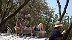 Turistas descansan en un banco bajo un paraguas frente a la colina de la Acrópolis durante una ola de calor en Atenas, el miércoles 9 de julio de 2025. 