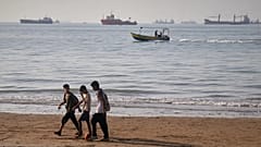 Kinder gehen am Ufer entlang, während ein Motorboot vor der Küste und Massengutfrachter in der Straße von Hormuz vorbeifahren, 27. April 2026