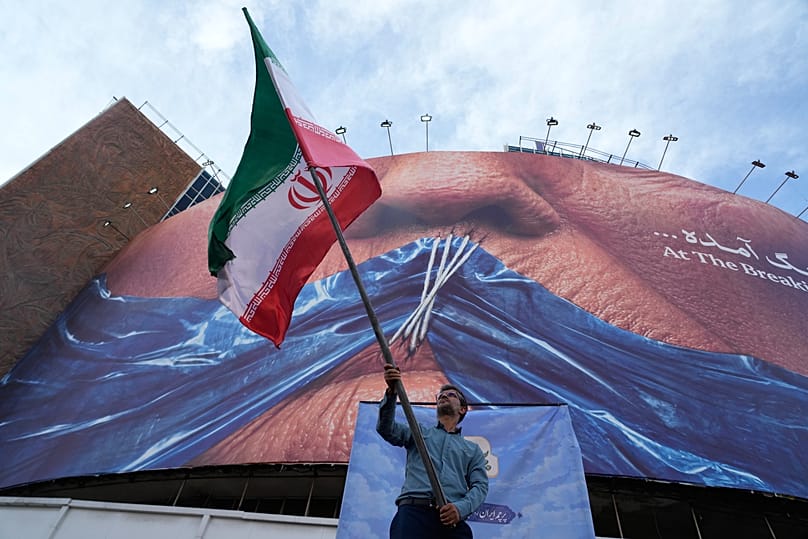 A man waves an Iranian flag under a billboard with graphic showing the sewn lips of US President Donald Trump in Tehran, 6 May, 2026