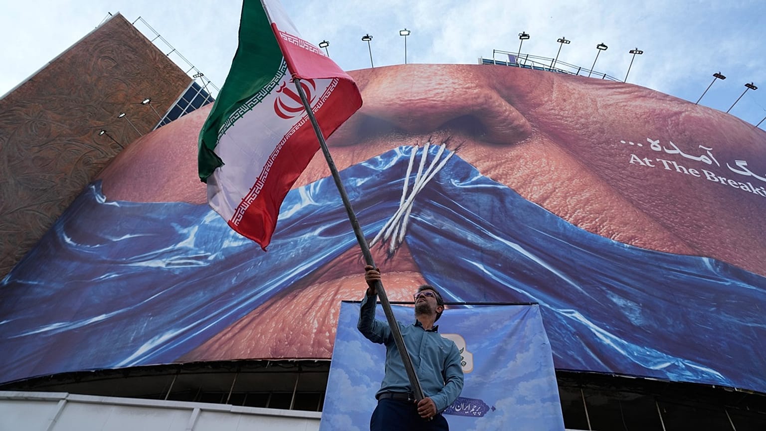 A man waves an Iranian flag under a billboard with graphic showing Strait of Hormuz and sewn lips of US President Donald Trump in downtown Tehran, Wednesday, May 6, 2026.