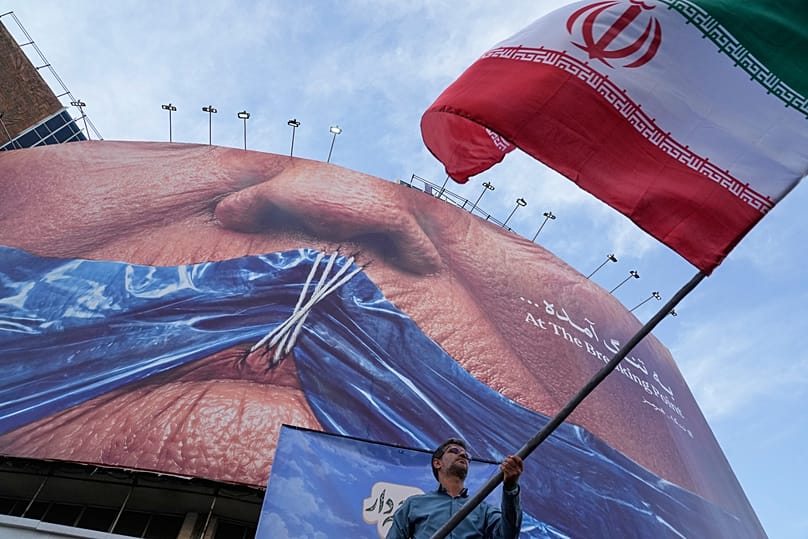 A man waves an Iranian flag under a billboard with graphic showing Strait of Hormuz and sewn lips of US President Donald Trump in downtown Tehran, 6 May 2026