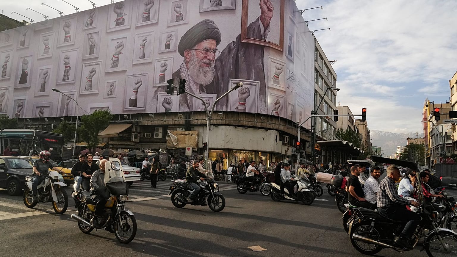Teherán belvárosában motorosok haladnak el egy óriásplakát mellett, amelyen a február 28-án amerikai és izraeli csapásokban megölt néhai legfelsőbb vezető Ali Hamenei látható.