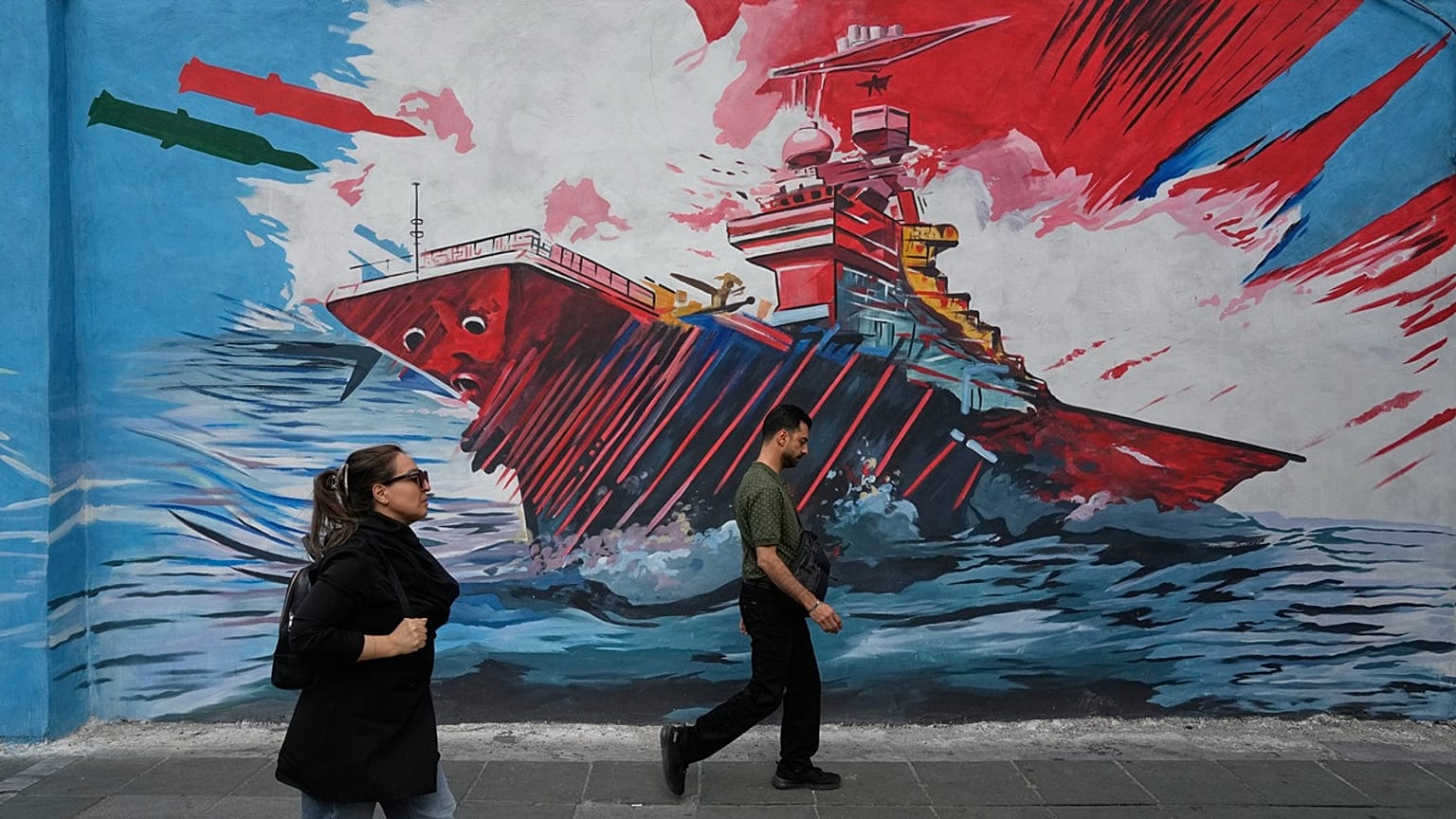 People walk past a mural depicting a U.S. aircraft carrier under missile attack in downtown Tehran, Iran, Sunday, May 17, 2026.