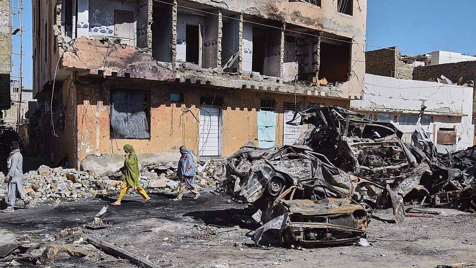 People walk past damaged cars and a building at the scene of Sunday's bomb explosion near a railway track in Quetta, 25 May, 2026