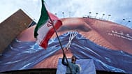 رجل يلوح بالعلم الإيراني في إطار حملة مؤيدة للحكومة، تحت لوحة إعلانية تحمل رسماً لمضيق هرمز وشفاه الرئيس الأمريكي المخيطة، في ساحة بوسط طهران، الأربعاء 6 مايو 2026.