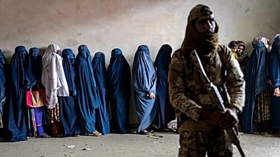 یک جنگجوی طالبان نگهبانی می‌دهد و زنان در کابل در ۲۳ مه ۲۰۲۳ منتظر دریافت جیره غذایی توزیع‌شده توسط یک گروه امدادرسان هستند