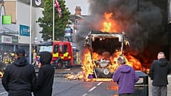 In East Belfast sehen Menschen zu, wie bei einem Protest ein Fahrzeug brennt, nach einer Messerattacke in Belfast am Dienstag, den neunten Juni 2026.