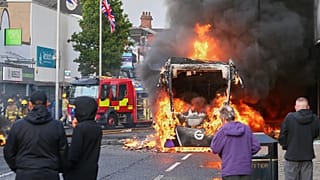 Alcune persone osservano un veicolo in fiamme durante una protesta a East Belfast, in seguito a un accoltellamento avvenuto a Belfast, martedì 9 giugno 2026.