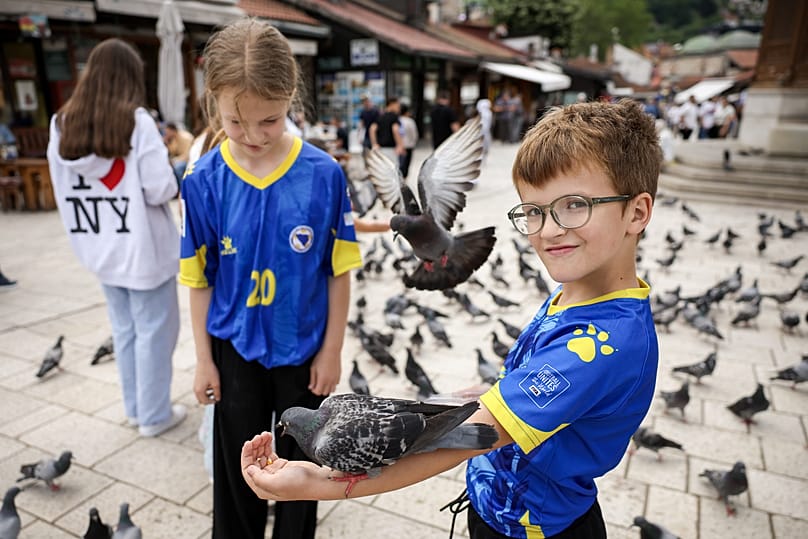 Bosna Milli Takımı formaları giyen çocuklar, 12 Haziran 2026’da Kanada-Bosna Dünya Kupası maçı öncesi Saraybosna’nın eski çarşısında güvercinlere yem veriyor