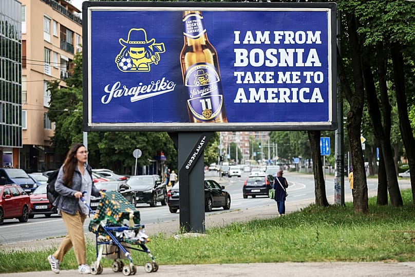 Bir kadın, 11 Haziran 2026’da Saraybosna’da Dubioza Kolektiv’in “I Am From Bosnia, Take Me to America” şarkısının sözlerinin yazılı olduğu bir reklam panosunun önünden geçiyor