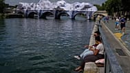 Persone sedute lungo la Senna accanto a “The Pont Neuf Cave”, installazione artistica gonfiabile dello street artist francese JR.