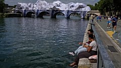 Persone sedute lungo la Senna accanto a “The Pont Neuf Cave”, installazione artistica gonfiabile dello street artist francese JR.