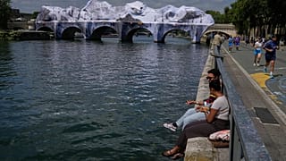 Des personnes sont assises le long de la Seine, à côté de « The Pont Neuf Cave », une installation artistique gonflable du street artiste français JR.