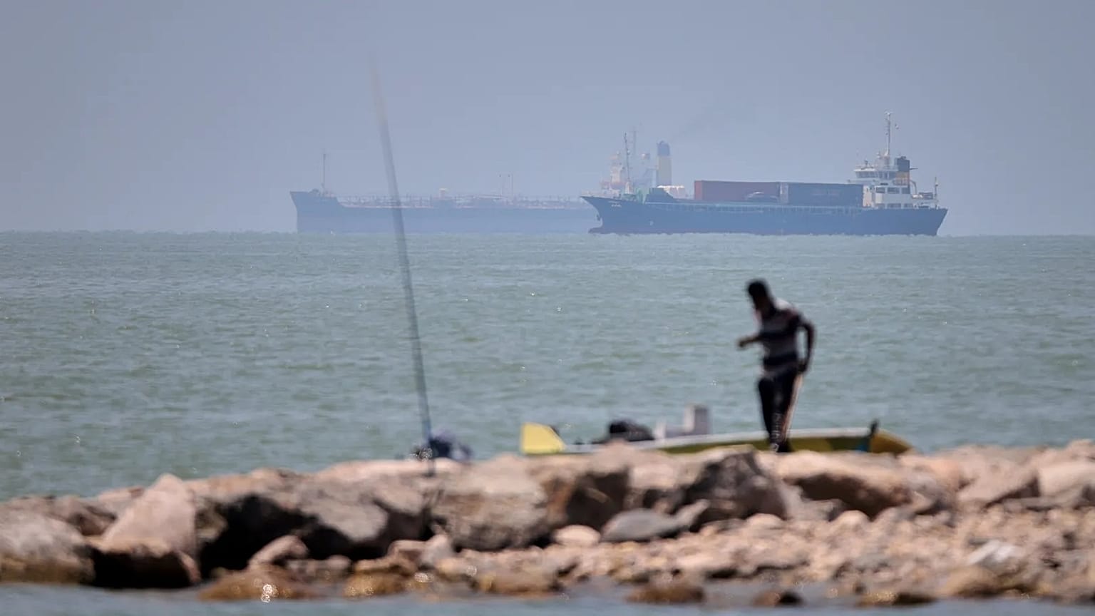 A man stands beside a fishing pole along the shore as cargo ships and commercial vessels are seen in the Strait of Hormuz off Bandar Abbas, Iran.