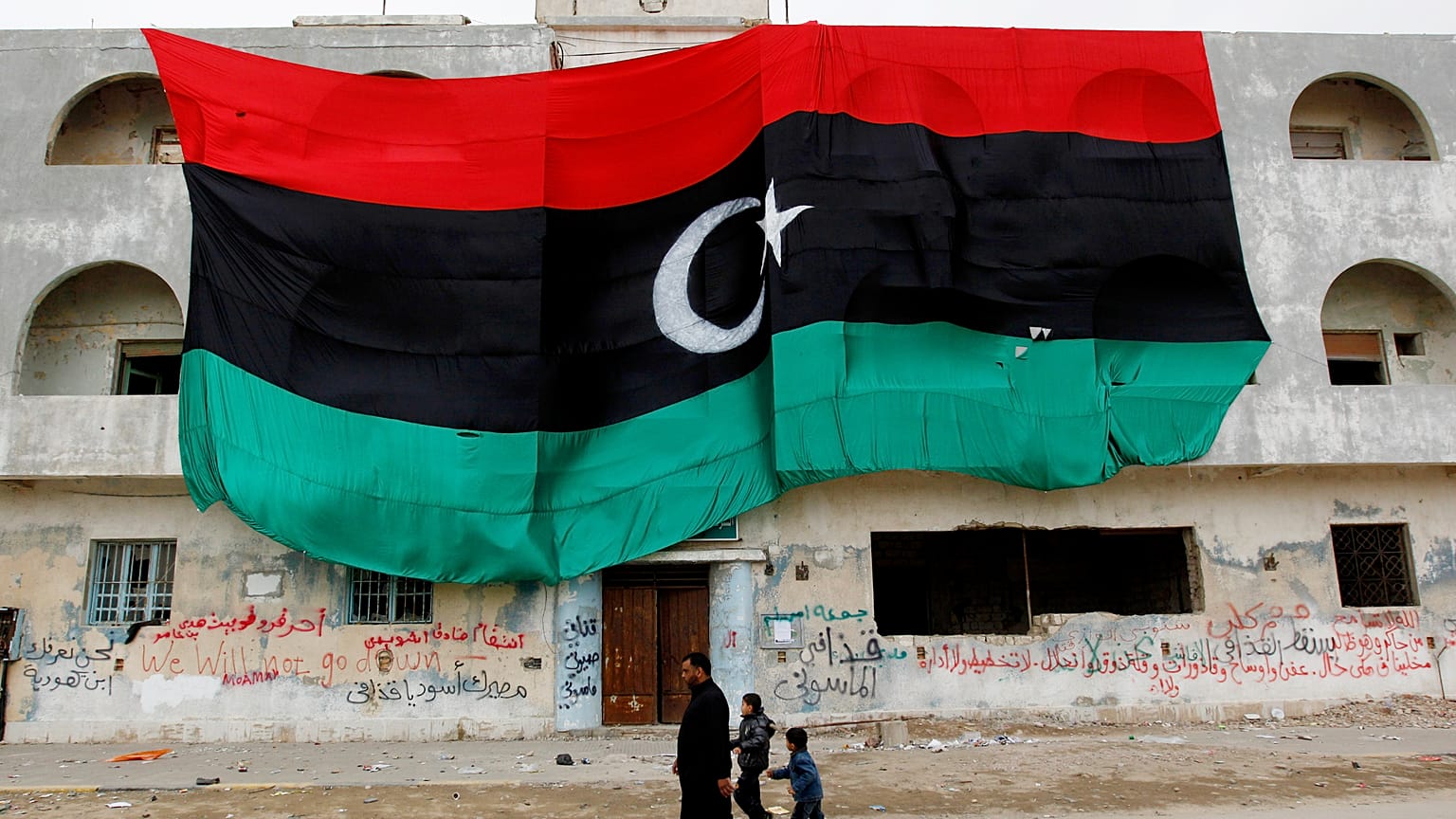 مواطنون ليبيون يمرون بمبنى يحمل العلم الليبي القديم الضخم، في بنغازي، شرق ليبيا، الجمعة 4 مارس 2011.
