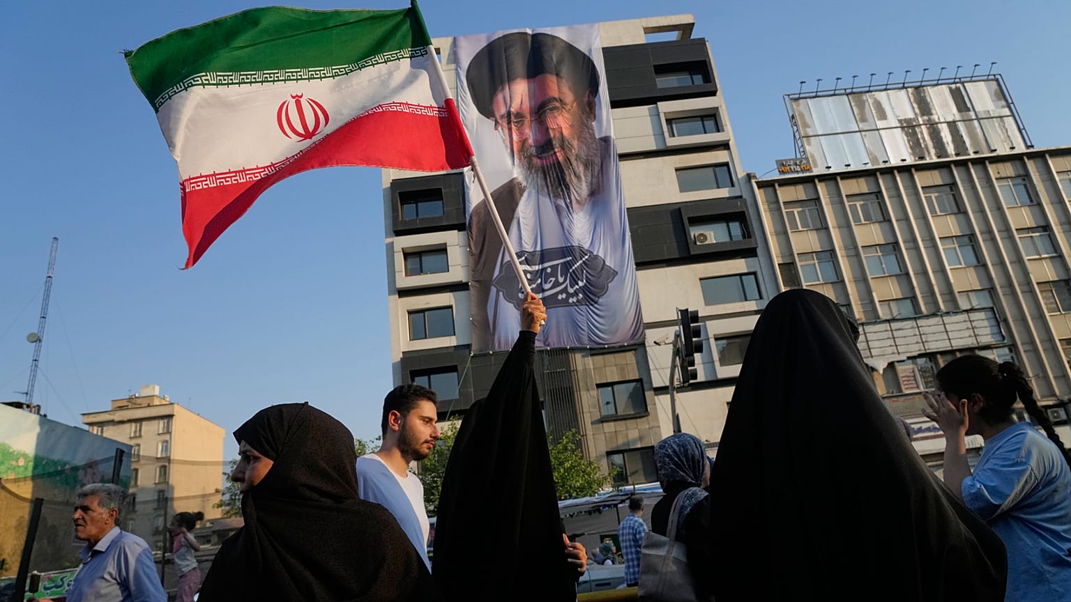 امرأة ترفع العلم الإيراني أمام لافتة تحمل صورة المرشد الأعلى آية الله مجتبى خامنئي خلال مهرجان شعبي احتفالاً بعيد الغدير، ذكرى تسمية النبي محمد صلى الله عليه وسلم لخليفته علي 