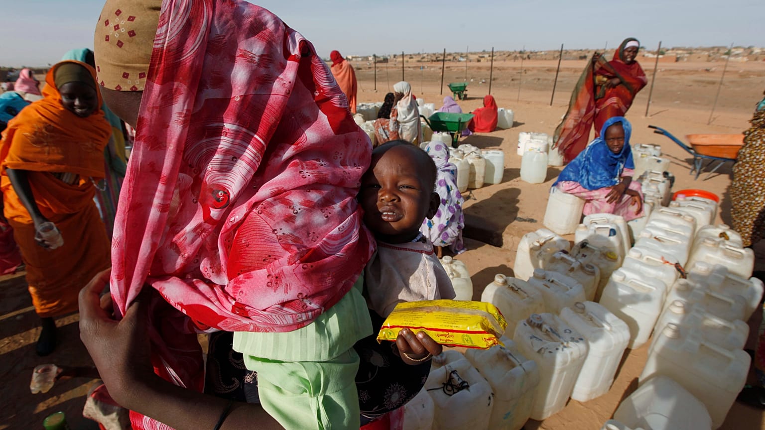 لاجئ سوداني يحمل طفلاً أثناء انتظاره لجمع إمدادات المياه في مخيم أبو شوك للاجئين خارج بلدة الفاشر بدارفور بالسودان