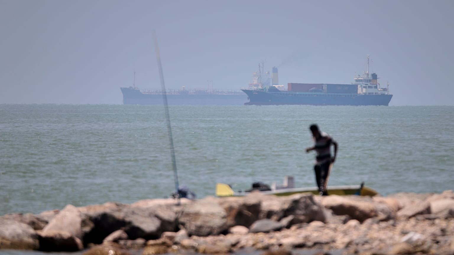 A man stands beside a fishing pole along the shore as cargo ships and commercial vessels are seen in the Strait of Hormuz off Bandar Abbas, Iran, Wednesday, June 17, 2026.