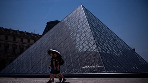 Turistas de chapéu-de-chuva caminham junto ao Museu do Louvre durante uma vaga de calor em Paris, sábado, 27 de junho de 2026.