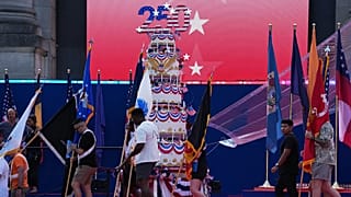 Persone sfilano con bandiere davanti a una torta gigante per festeggiare durante l’evento America 250 a Bruxelles.
