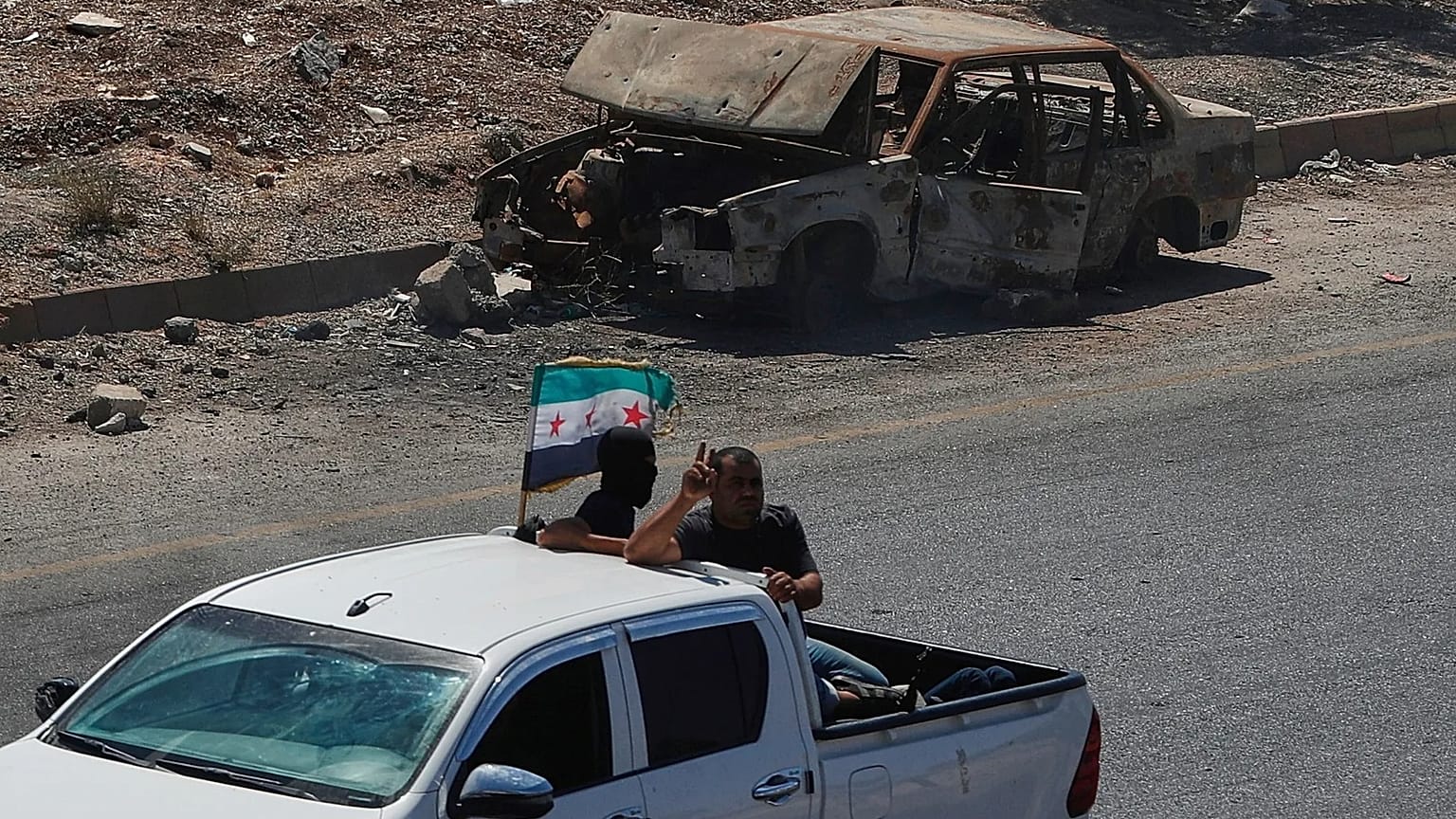  مقاتلون يستقلون الجزء الخلفي من شاحنة صغيرة ترفع العلم السوري، أثناء مرورهم بجوار سيارة محترقة على الطريق السريع الواصل بين درعا ودمشق في سوريا