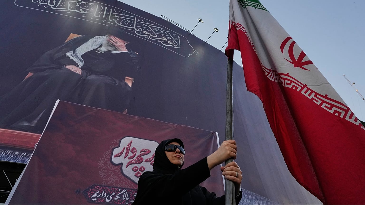 تحمل امرأة مؤيدة للحكومة العلم الإيراني تحت لافتة تحمل صورة المرشد الأعلى الراحل، علي خامنئي، في أحد ميادين وسط العاصمة طهران، إيران، الثلاثاء 30 يونيو/حزيران 2026.
