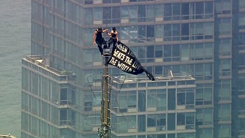 Two people stand on the tip of the antenna of the Empire State Building in New York while holding a banner, 1 July, 2026
