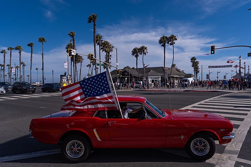 A vintage Ford Mustang carrying a U.S. flag passes through an intersection in Huntington Beach, Calif., Friday, July 3, 2026, ahead of the 250th anniversary.