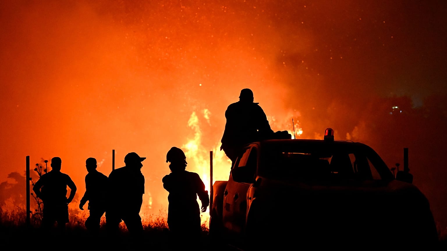 Bomberos y otras personas se recortan sobre un incendio forestal en las afueras de la ciudad norteña de Salónica, Grecia, el sábado cuatro de julio de 2026.