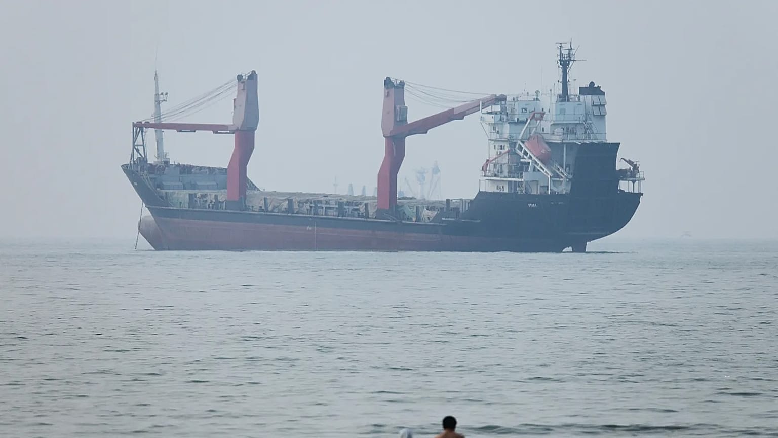 A group of people stands in shallow water as a cargo ship appears anchored in the Strait of Hormuz off Bandar Abbas, Iran, Tuesday, June 30, 2026.