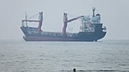 A group of people stands in shallow water as a cargo ship appears anchored in the Strait of Hormuz off Bandar Abbas, Iran, Tuesday, June 30, 2026.