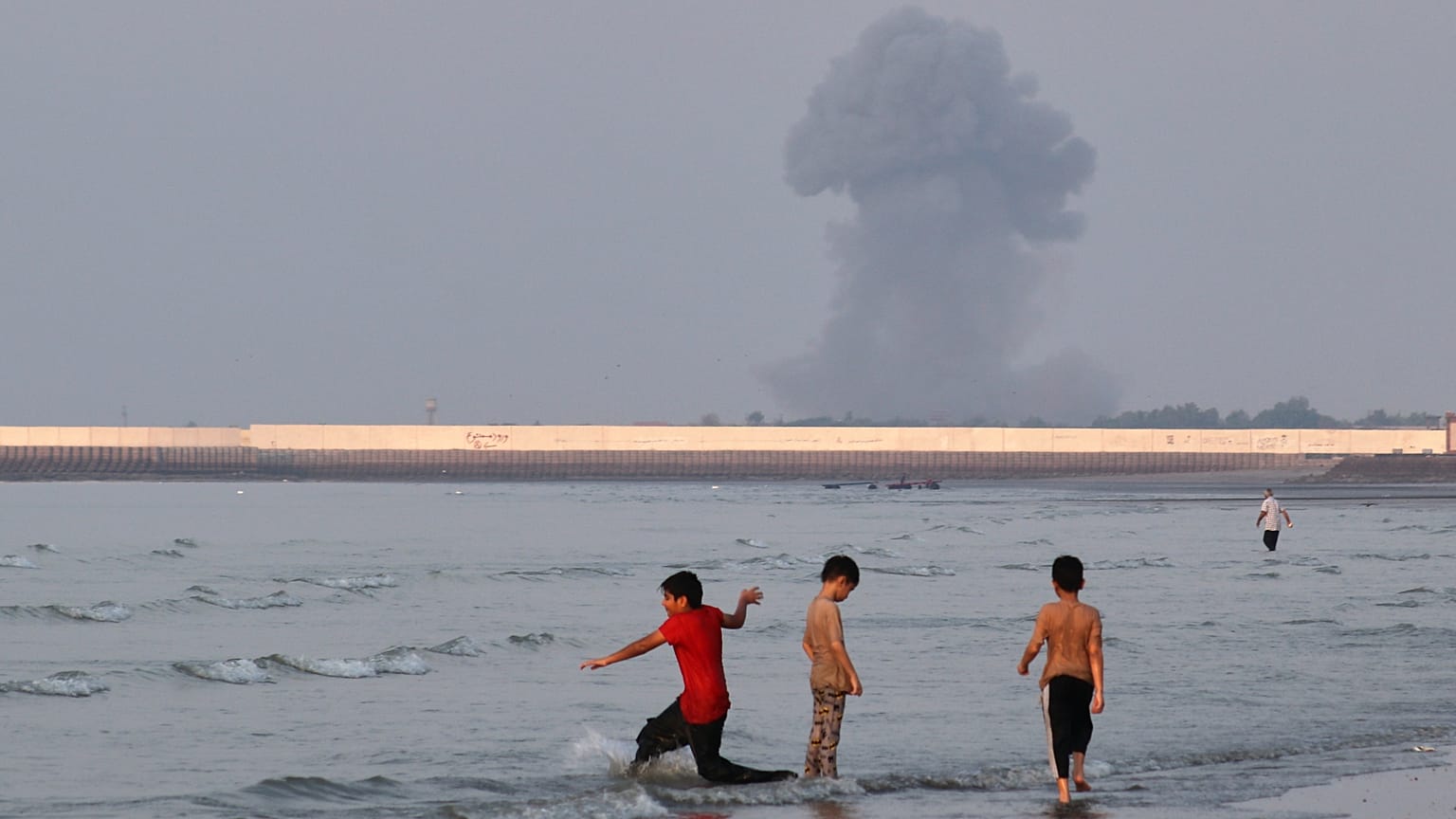 Three boys play in the shallow waters of the Strait of Hormuz, as a plume of smoke rises from an explosion in the background, off Bandar Abbas, Iran, Monday, July 13, 2026. 