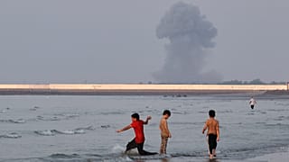 Three boys play in the shallow waters of the Strait of Hormuz, as a plume of smoke rises from an explosion in the background, off Bandar Abbas, Iran, Monday, July 13, 2026. 
