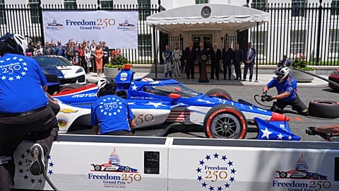 A pit crew demonstrates a tire change as President Donald Trump, in background third from left
