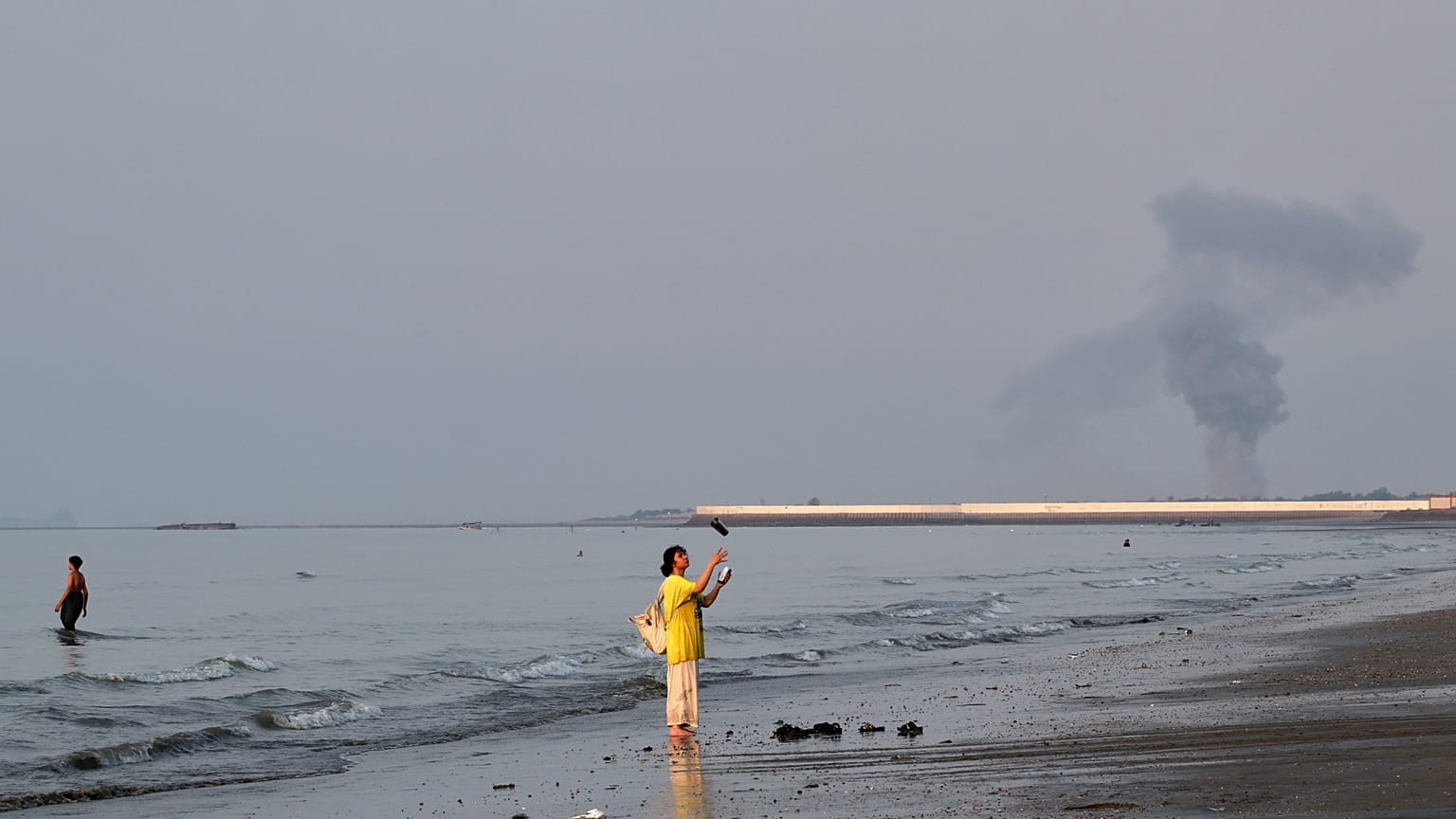 A woman stands at the water's edge along the Strait of Hormuz as a plume of smoke rises in the background following an explosion, off Bandar Abbas, Iran, Monday, July 13, 2026
