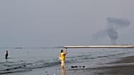 A woman stands at the water's edge along the Strait of Hormuz as a plume of smoke rises in the background following an explosion, off Bandar Abbas, Iran, Monday, July 13, 2026