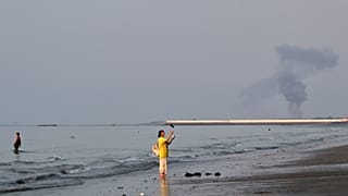 A woman stands at the water's edge along the Strait of Hormuz as a plume of smoke rises in the background following an explosion, off Bandar Abbas, Iran, Monday, July 13, 2026
