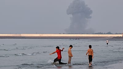 Three boys play in the shallow waters of the Strait of Hormuz as a plume of smoke rises from an explosion in the background off Bandar Abbas, Iran, 13 July 2026
